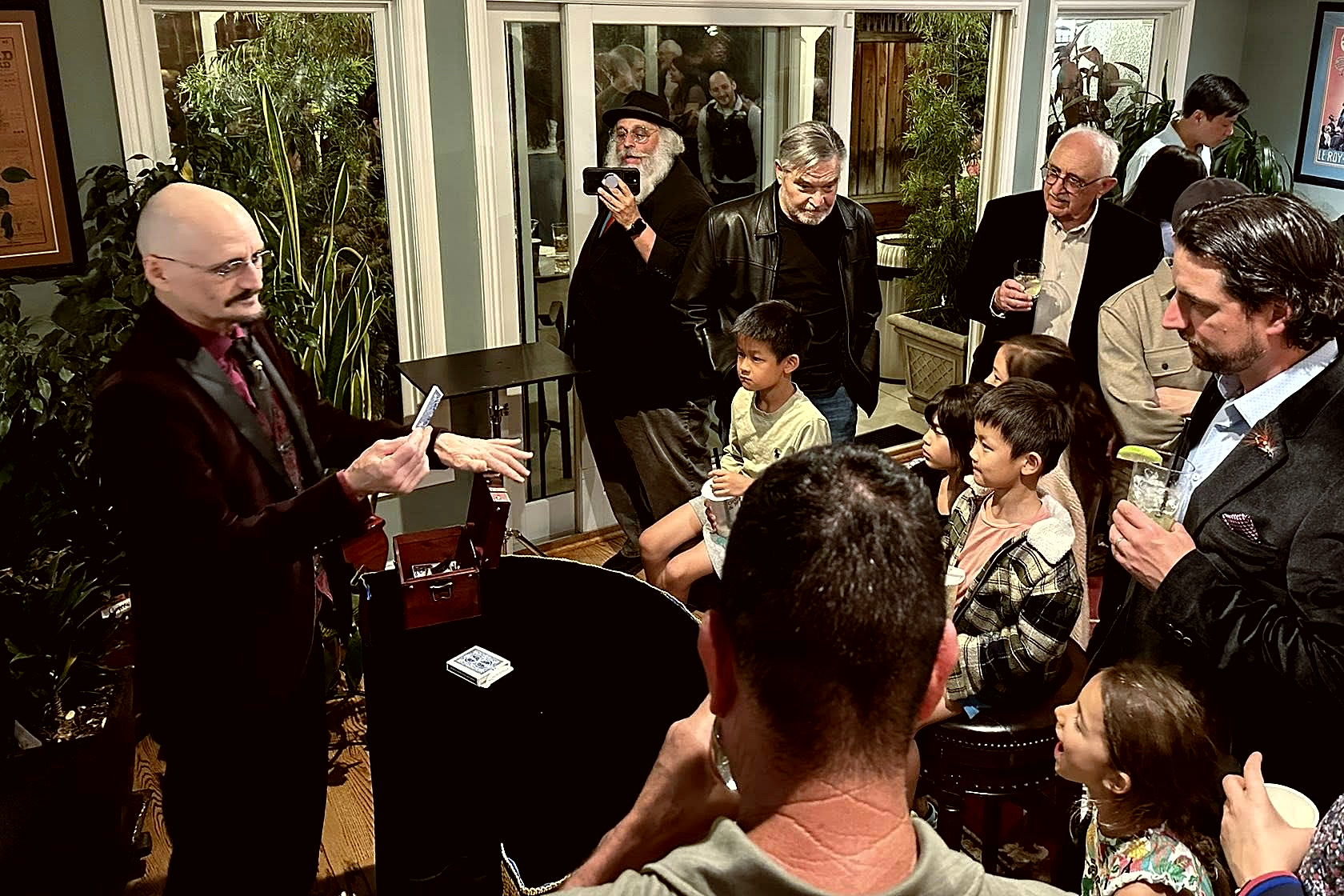 Ace Miles working close-up magic to a packed room, card held out toward a crowd of adults and children standing shoulder to shoulder, with vintage magic posters on the walls.