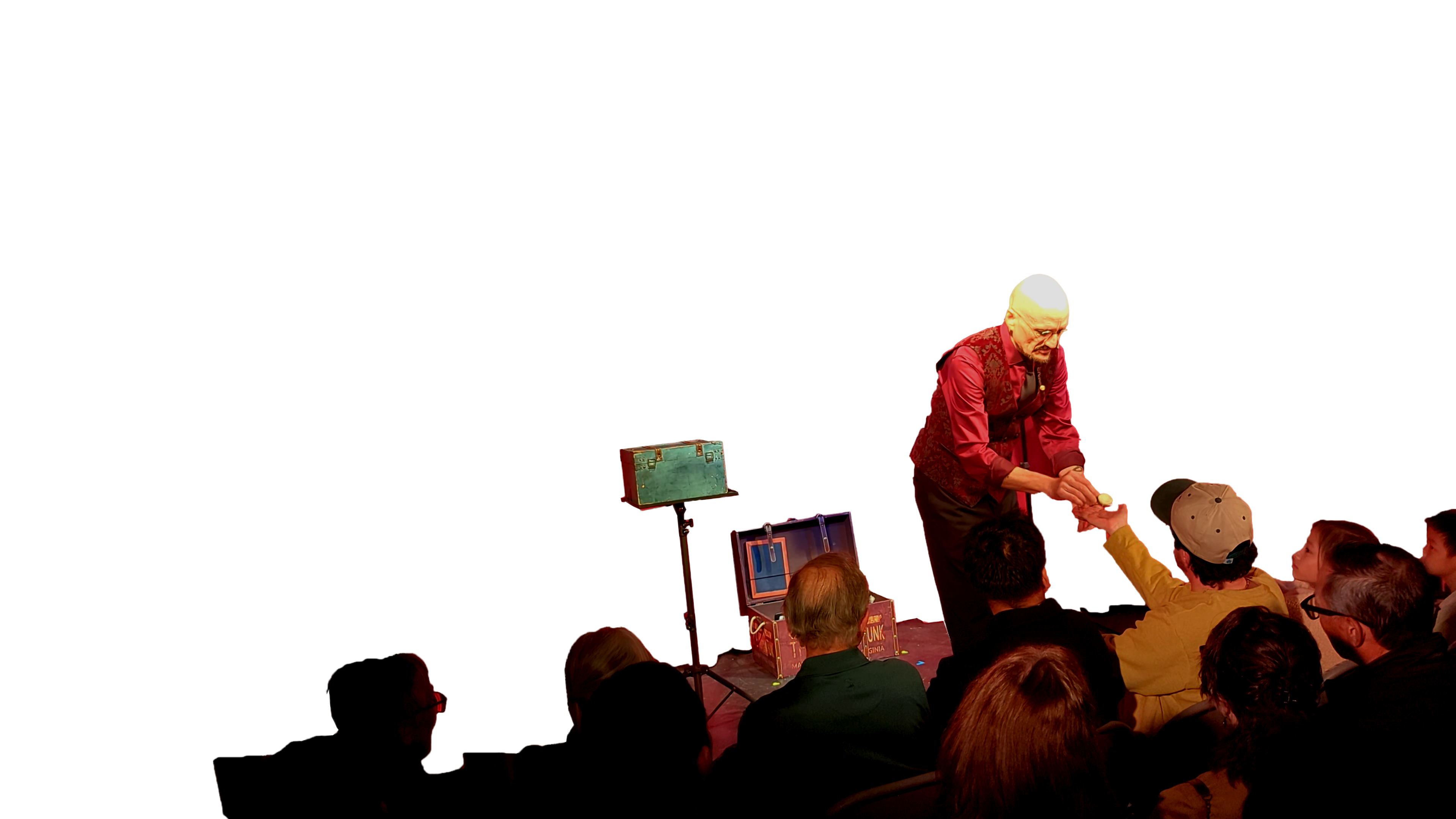 Ace Miles on a theater stage in front of a deep red velvet curtain, leaning down to hand a small object to a man in the front row, with the audience watching in silhouette.