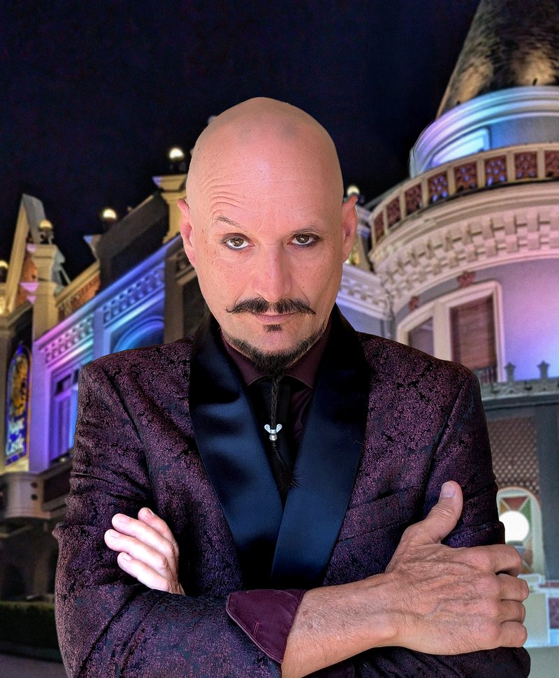 Ace Miles, arms crossed, in a dark purple brocade dinner jacket, standing in front of the Magic Castle's lit turret in Hollywood at night, 2025.