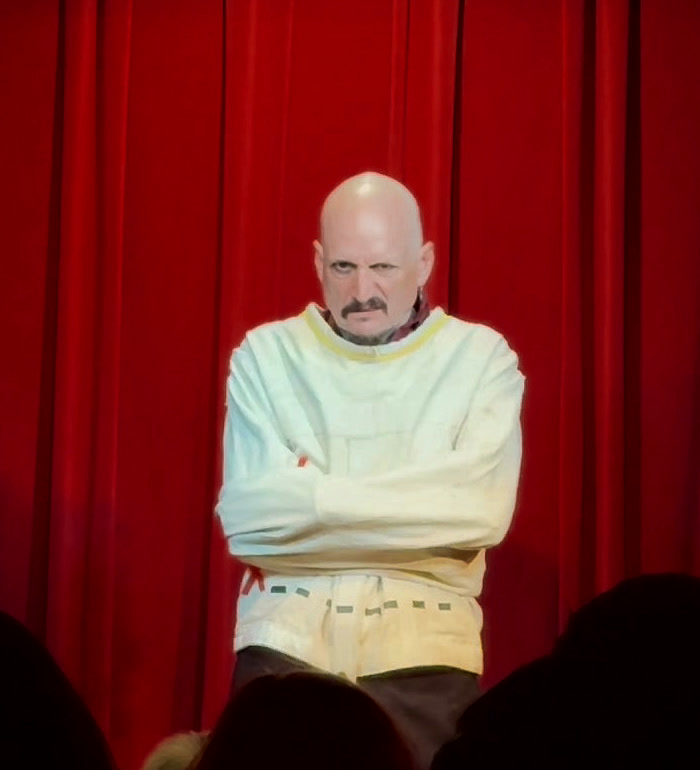 Ace Miles on stage in a white straitjacket, arms buckled across his chest, standing still in front of a red curtain with the audience in silhouette.